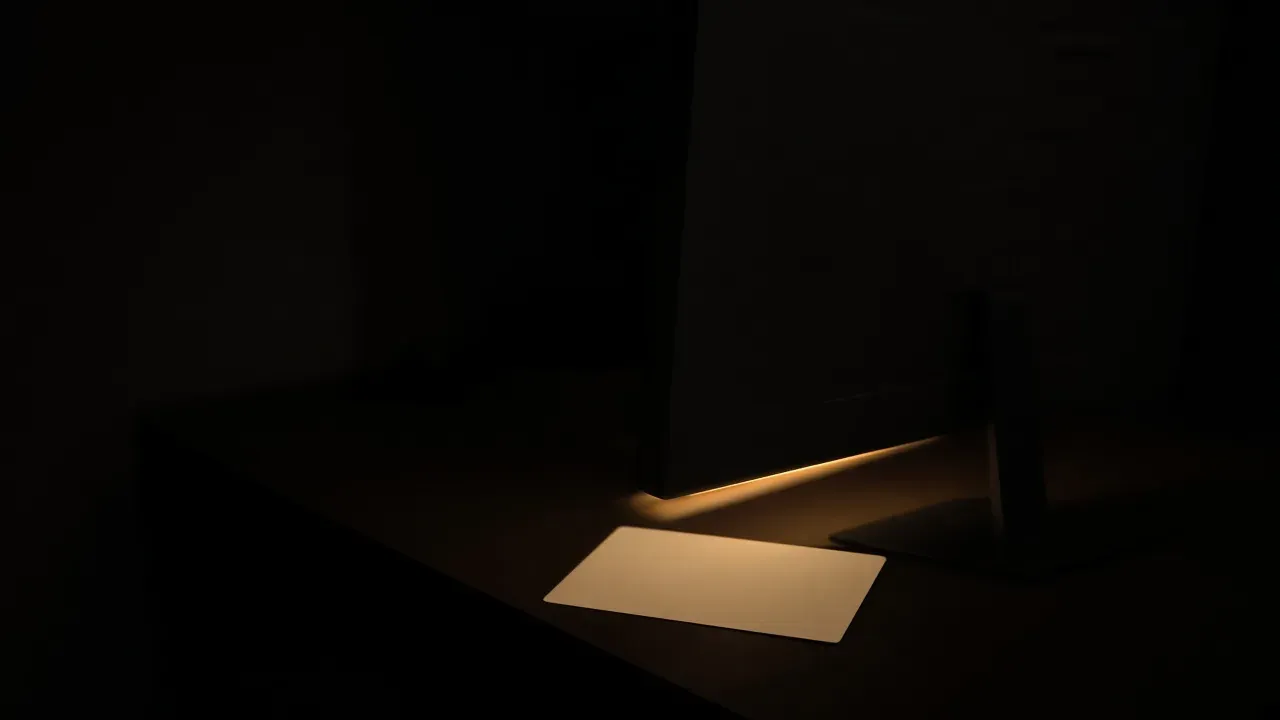The back of a dark monitor on a desk, with a warm amber glow along its lower edge lighting a blank card